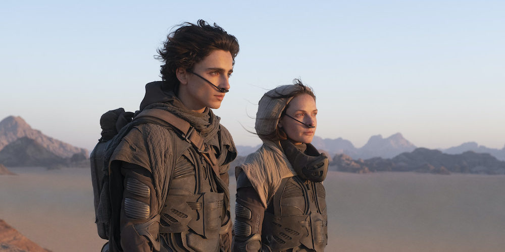 Image of Timothee Chalamet playing Paul Atreides and Rebecca Ferguson playing Jessica in the film Dune. They are both dressed in brown clothing staring into the distance in front of a backdrop of desert and mountains