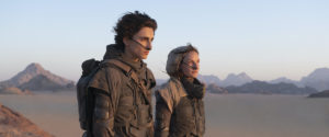 Image of Timothee Chalamet playing Paul Atreides and Rebecca Ferguson playing Jessica in the film Dune. They are both dressed in brown clothing staring into the distance in front of a backdrop of desert and mountains