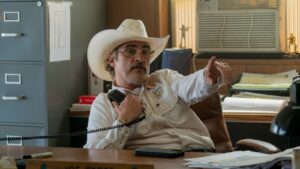 A bearded, bespectacled sheriff sits at his desk, pointing and giving instructions.