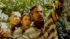 Three men in prison attire crouching in a wood.
