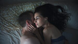 A man and woman lying in bed, embracing as moonlight illuminates the woman’s face while the man sleeps.