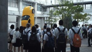 Japanese students gathered in a courtyard looking at a yellow sports car standing vertically on its nose.