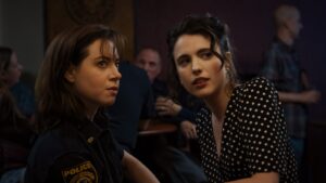 Aubrey Plaza in police uniform and Margaret Qualley in a black dress sitting together at a bar with curious expressions.
