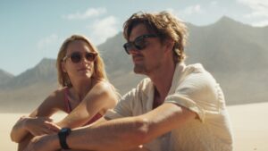 Sam Riley as a young man sitting with a woman on a sandy beach with mountains in the background.
