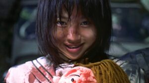 A young Japanese girl covered in blood smiles holding a teddy bear.