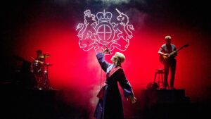 Rosamund Pike dressed as a judge stands on a stage holding a microphone about to sing.