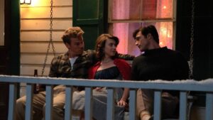 Two young men and a young woman in 1950s dress sit on the bench of a wooden farmhouse porch.