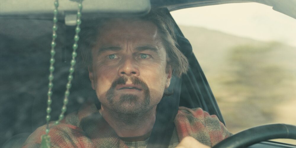 Leonardo DiCaprio as a revolutionary character driving a worn-out car, nervously glancing at the rear-view mirror.