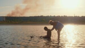Two people bathe in a river at sunset as smoke rises from a bombed Ukrainian village in the background.