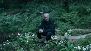 Daniel Day-Lewis sits on a log in a wooded area.