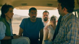 A group of people sit in the back of a van looking worried, including a woman in a wedding dress