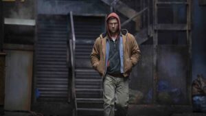 Glen Powell, wearing a hoody and looking moody, walks toward camera